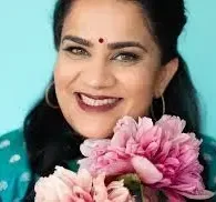 Immigrant Indian comedian Zarna Garg holindg pink flowers in a green background with a traditional bindi on her head.